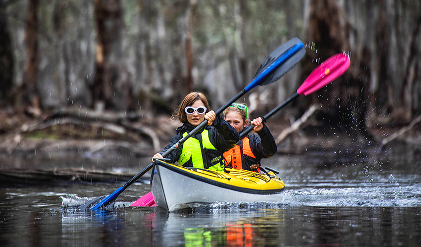 A lady and a girl paddling in a kayak on a wetland with redgum forest behind them