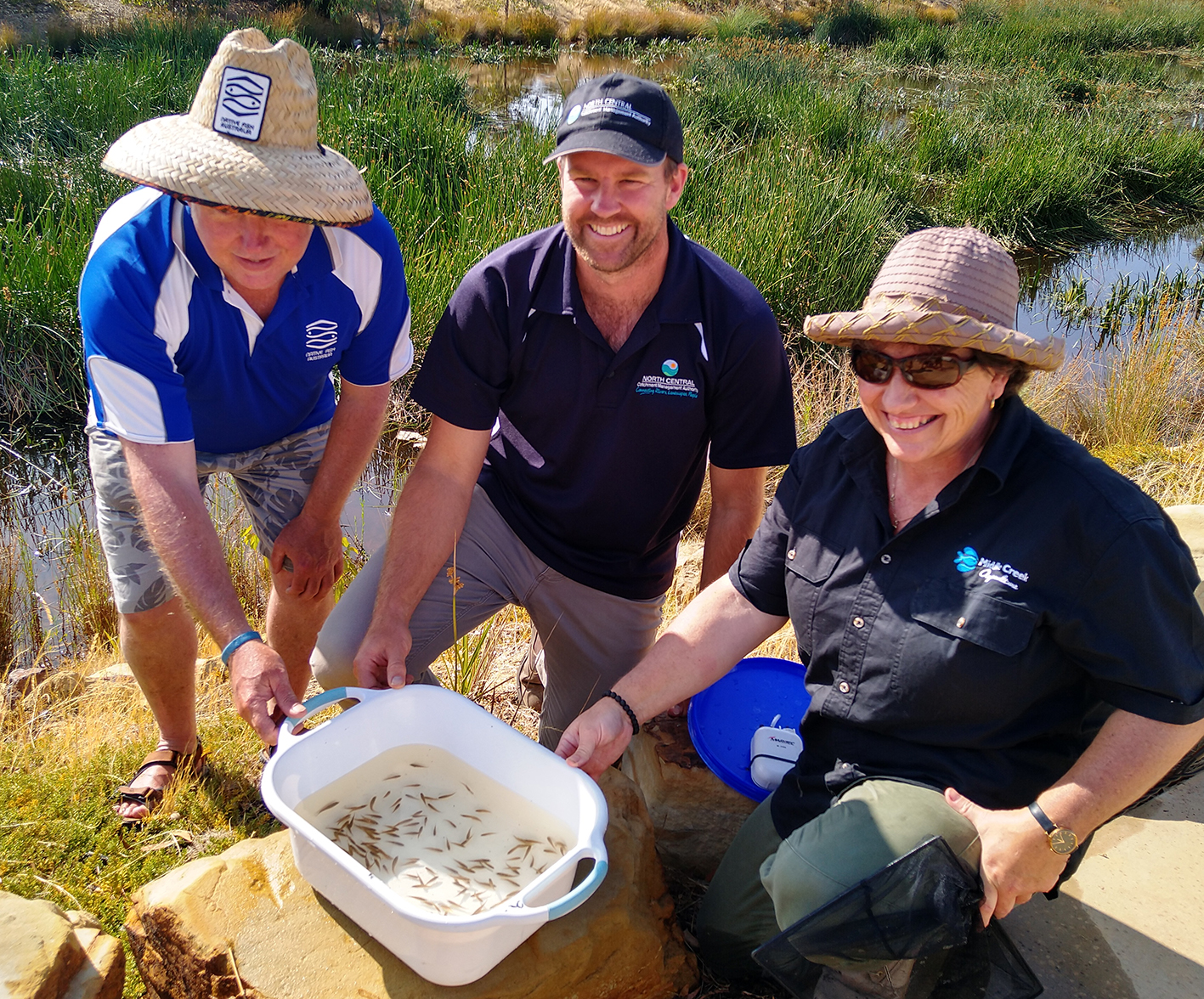 two men and a woman squatting around a bucket of small fish with a wetland in the background