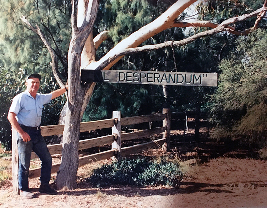 S middle aged man standing next to a tree, a fence, and a property sign that reads: "Desperandum"