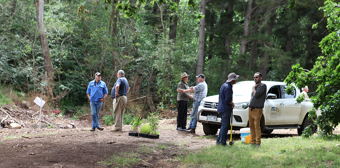 Six men in the middle ground in groups of 2, standing around talking to each other. One pair is leaning on a white North Central CMA ute. In the background is a forest