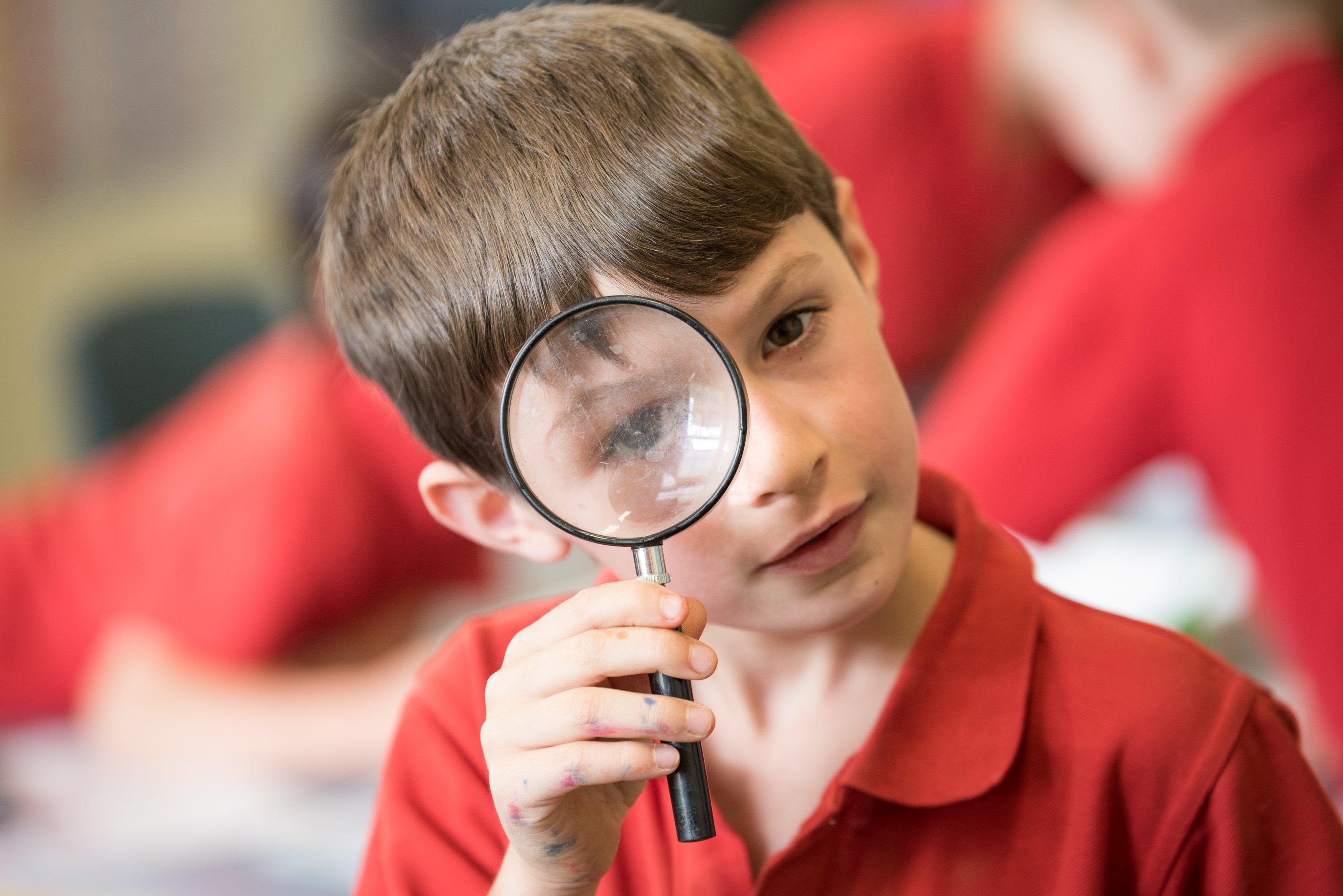 Primary school aged boy wearing a red shirt holding a magnifying glass close to his right eye.