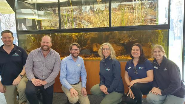 Three men and three women - all CMA staff - kneel down in front of an aquarium that includes a open-air section above the water for reeds and sticks 