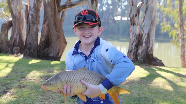 Young boy in a blue shirt and cap standing on the banks of a creel holding a carp and smiling.