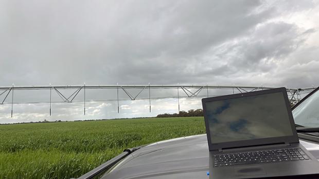 Laptop sits on a grey car bonnet in the foreground. Is the background is an irrigation sprayer over a green crop
