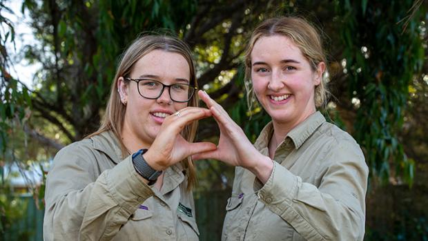 Two young women in khaki shirts holding their hands together to make a Landcare sign, like the map of Austrakia. 