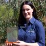 Tess with dark hair and a blue shirt stands in front of a young gum tree holding the Landcare Award trophy