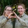Two young women in khaki shirts holding their hands together to make a Landcare sign, like the map of Austrakia. 